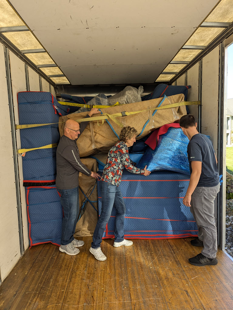 REPLACE: Ged and Clay working alongside a customer inside the AAGGO truck, securing a load with moving blankets and straps